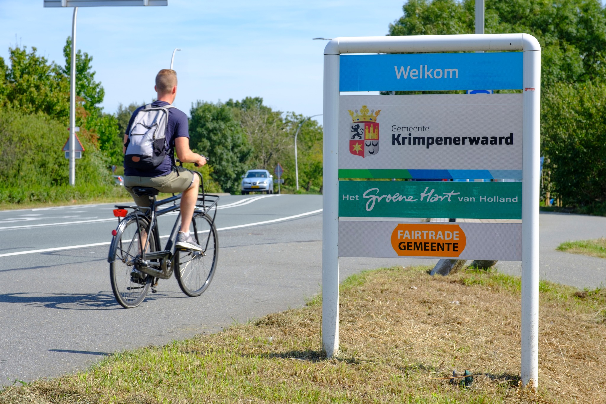 een man die op een fiets op een weg rijdt en daarbij het bord passeert met de tekst: Welkom in de Gemeente Krimpenerwaard.